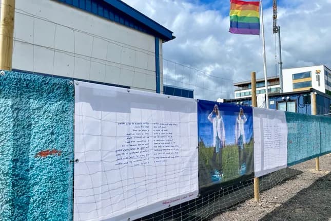 En rad affischer på ett staket med en regnbågsflagga i bakgrunden.