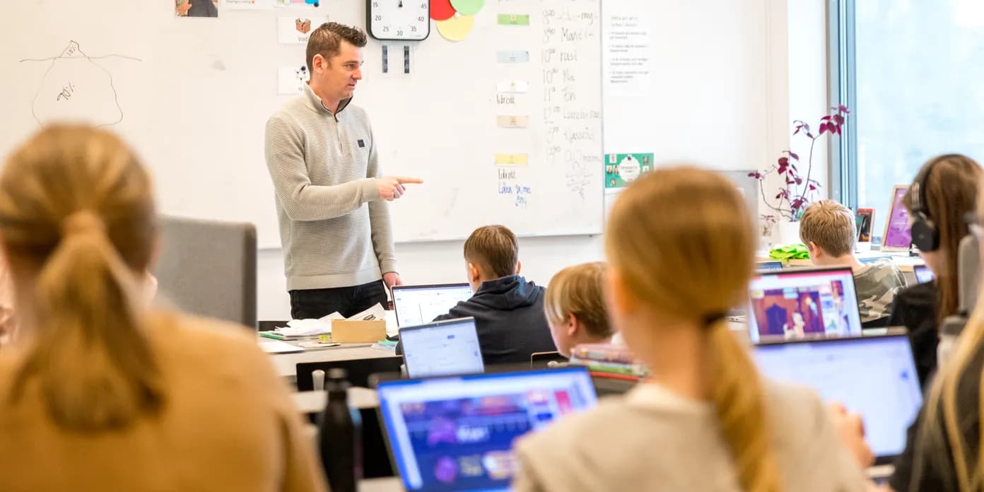 Mies seisoo luokkahuoneen edessä, joka on täynnä oppilaita.