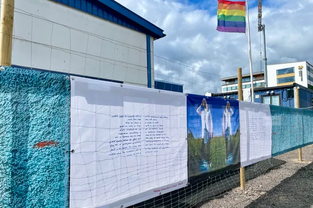 En rad affischer på ett staket med en regnbågsflagga i bakgrunden.